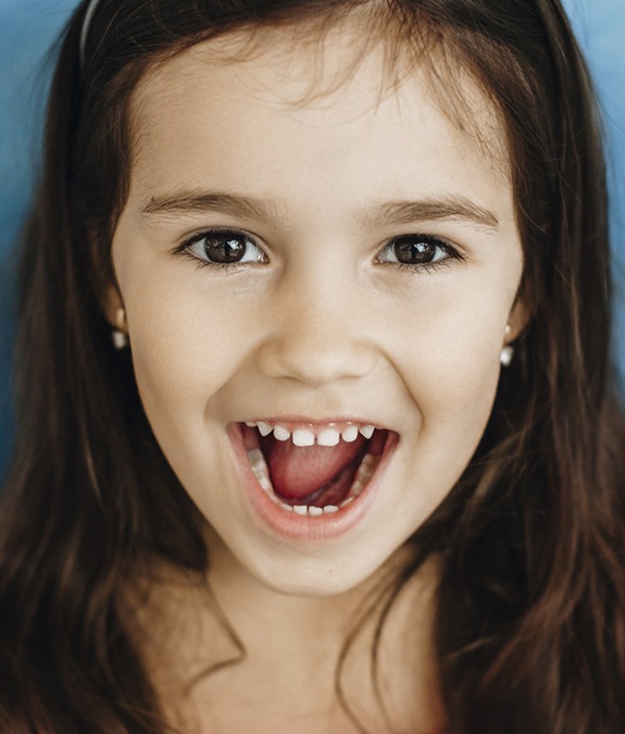A little girl with long, dark hair smiling with her mouth open to show off her healthy set of teeth