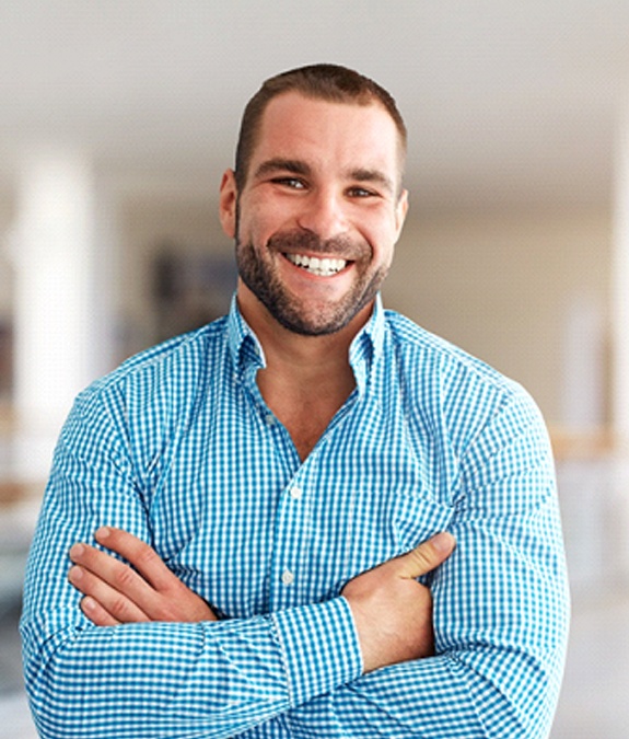 A younger man wearing a button-down shirt and folding his arms across his chest smiles after learning he’s eligible for a dental bridge